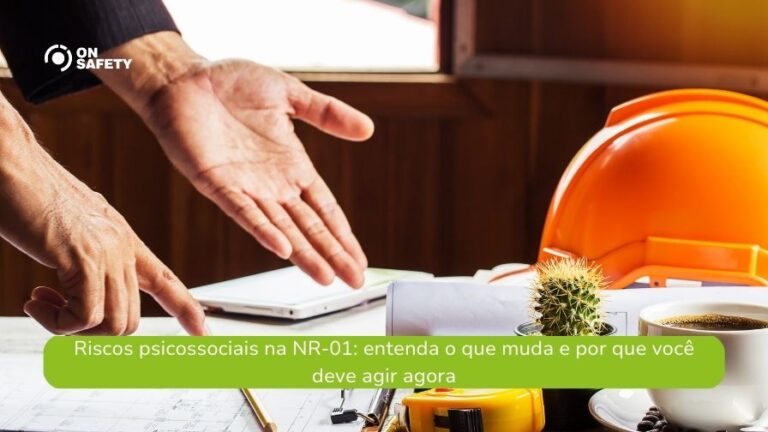 Em uma mesa de escritório, vemos objetos relacionados ao ambiente de trabalho, como capacete de segurança, xícara de café, fita métrica e documentos. Um pequeno cacto em vaso está em destaque ao centro. Mãos de uma pessoa gesticulam acima dos papéis, sugerindo uma conversa ou discussão remetendo à NR 01 e riscos psicossociais. O logo da OnSafety está no canto superior esquerdo. Uma faixa verde na parte inferior contém o texto: "Riscos psicossociais na NR-01: entenda o que muda e por que você deve agir agora".