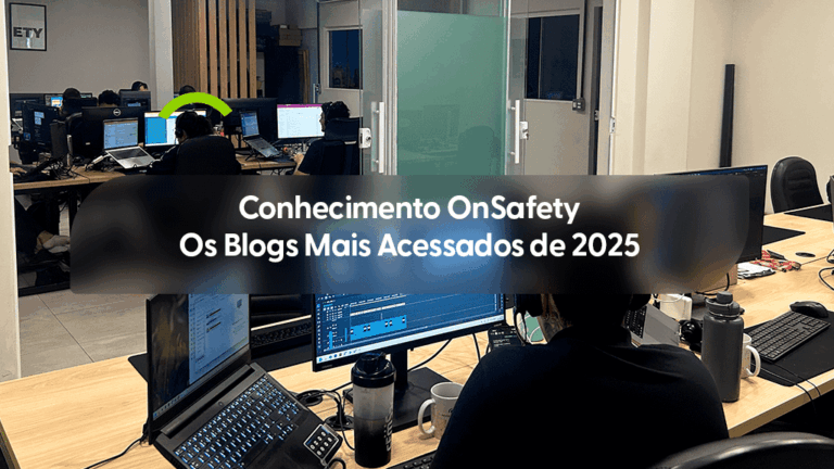 Vista interna de um escritório contemporâneo onde diversos profissionais trabalham em mesas equipadas com notebooks e múltiplos monitores. Em primeiro plano, a parte posterior de um colaborador focado em suas telas de trabalho. Uma forma circular verde estilizada aparece acima de um dos monitores ao fundo. Sobre a imagem, uma caixa de texto cinza transparente contém o título: "Conhecimento OnSafety Os Blogs Mais Acessados de 2025".
