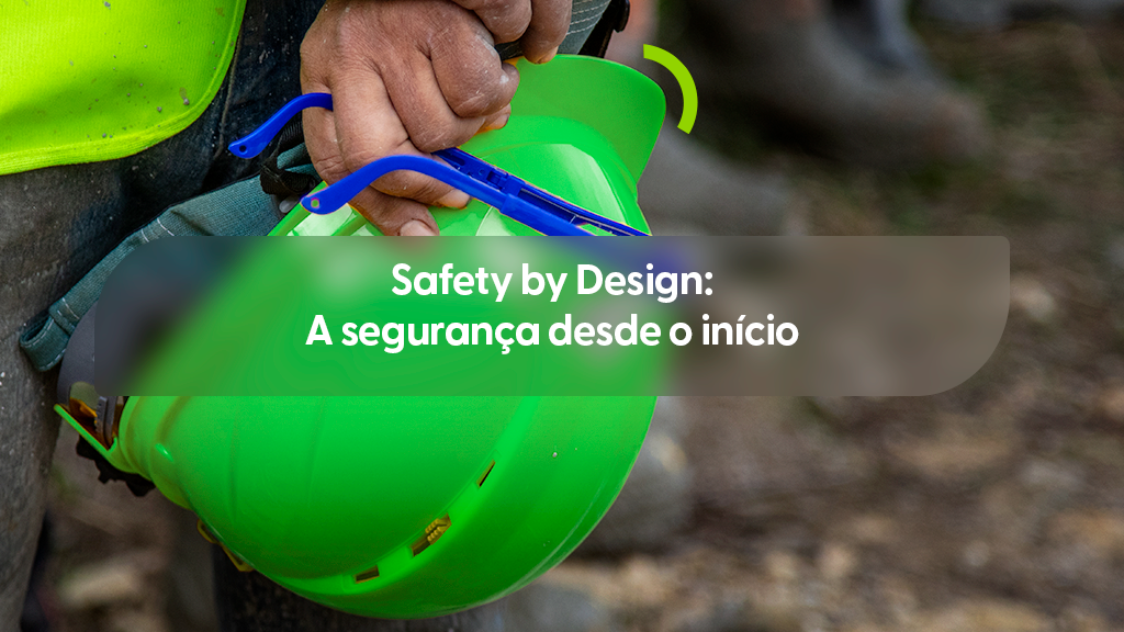 Imagem focada na mão de um trabalhador segurando um capacete de segurança verde limão e um par de óculos de proteção com armação azul. O profissional veste um colete refletivo verde e calça jeans. O fundo mostra um terreno de terra desfocado. Sobre a imagem, há uma caixa de texto cinza transparente com o título "Safety by Design: A segurança desde o início" e um pequeno detalhe em arco verde acima do capacete.